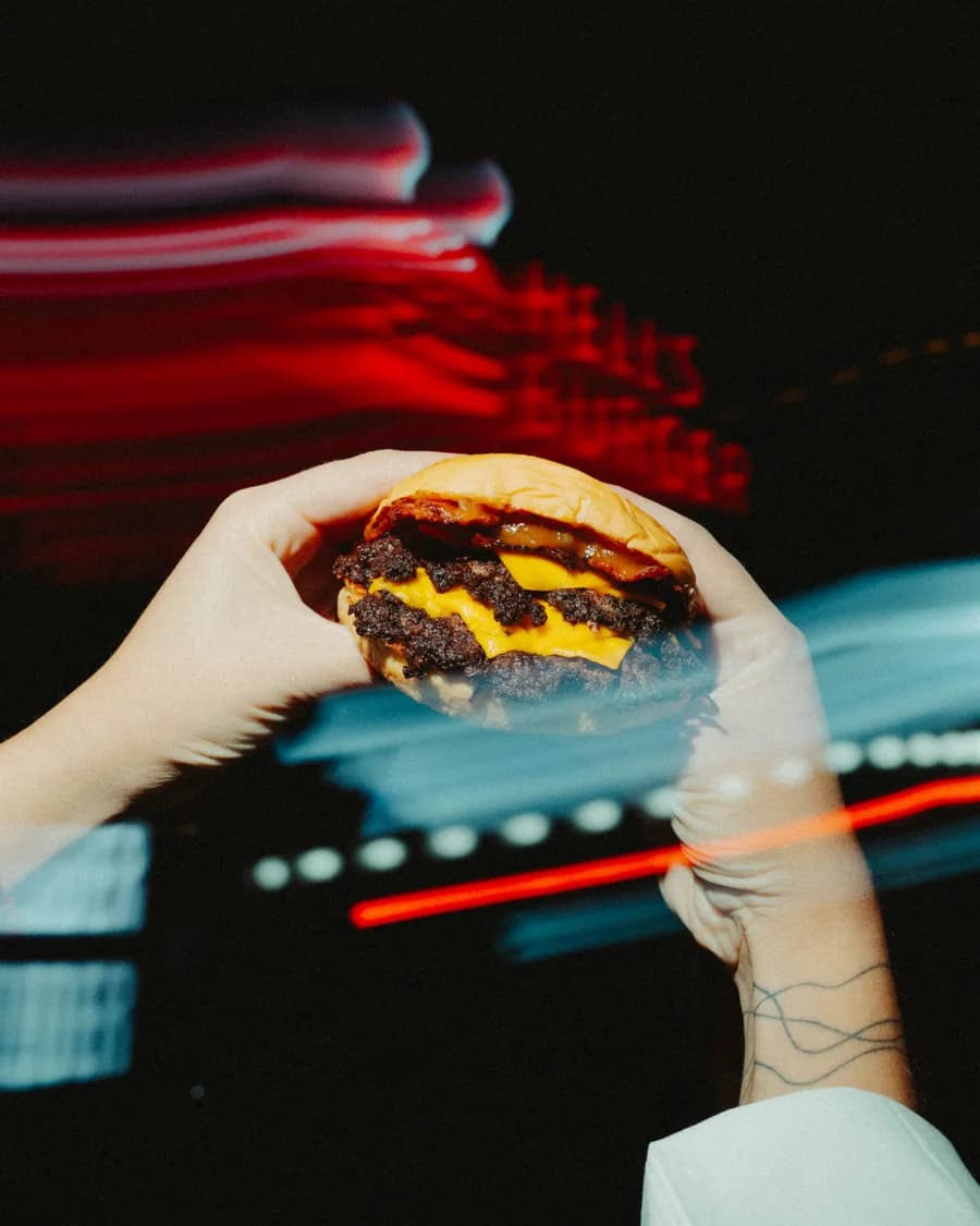 Dos manos sosteniendo una hamburguesa con cheddar frente a las luces de la calle